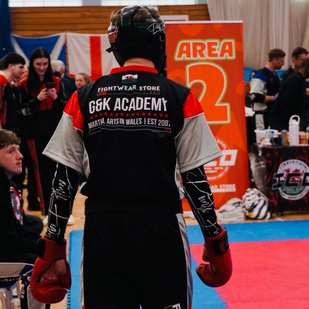 A person wearing a black, white, and red fight apparel uniform with 'G&K ACADEMY' and 'FIGHTWEAR STORE' printed on the back, standing on a blue and red mat in an indoor sports hall. Wearing Compression Sleeve Elbow Pads.